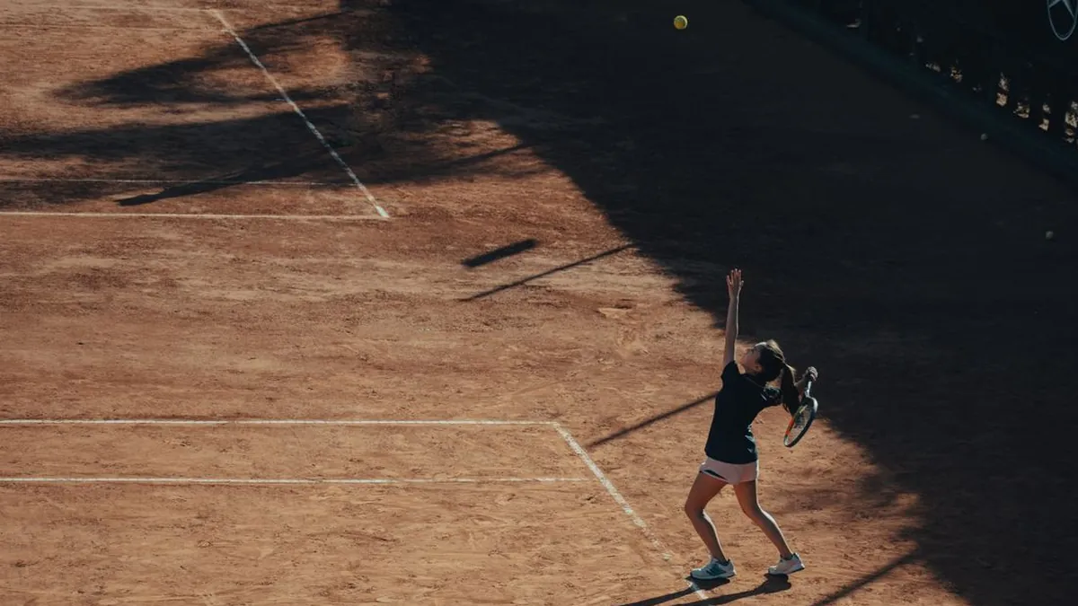 Joueuse de tennis au service, bras tendu vers la balle lancée en l'air, sur un court en terre battue